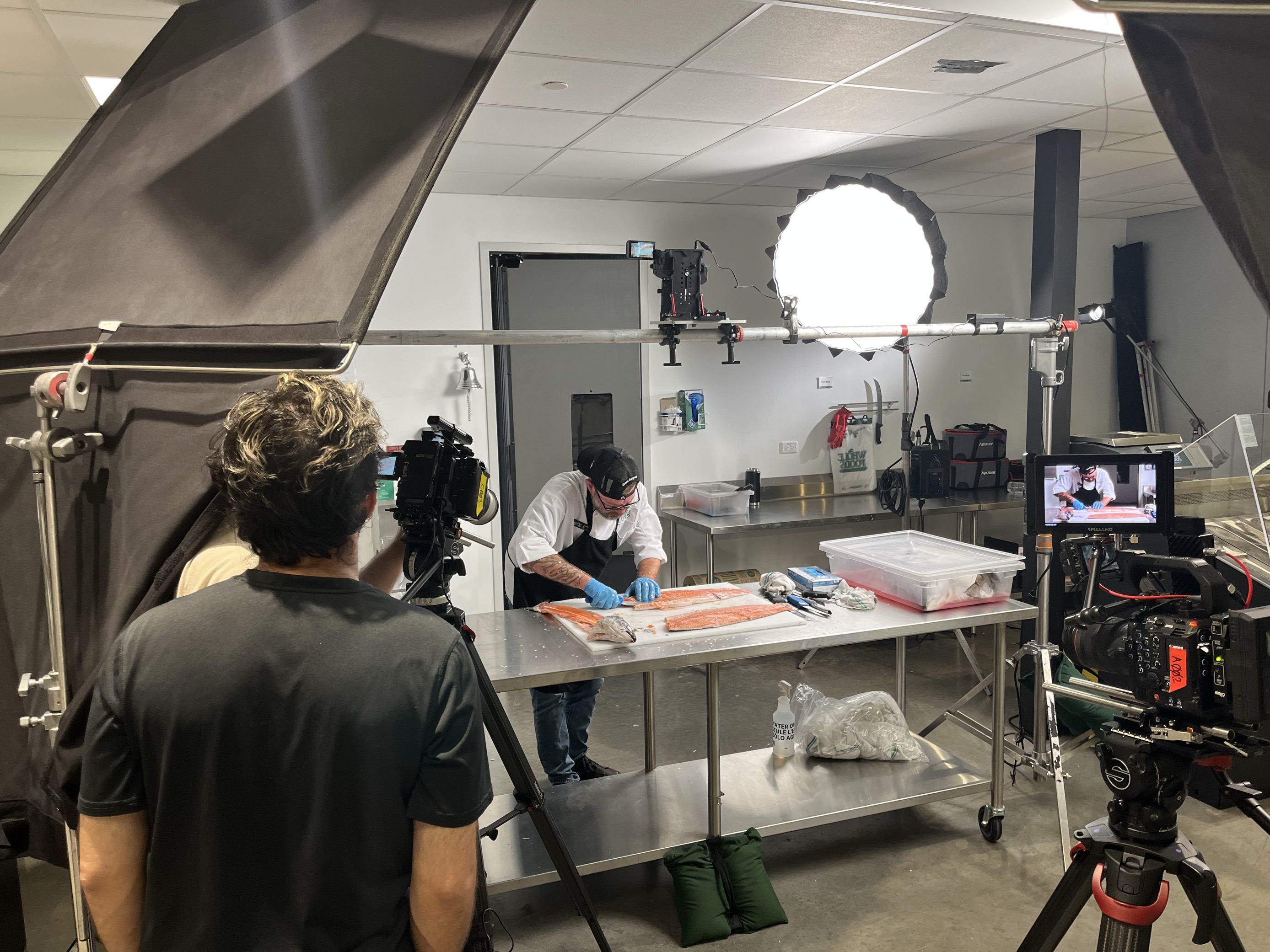 Behind-the-scenes photo of a chef filleting salmon on a stainless steel table while a film crew records the demonstration for the apprentice program's training video.
